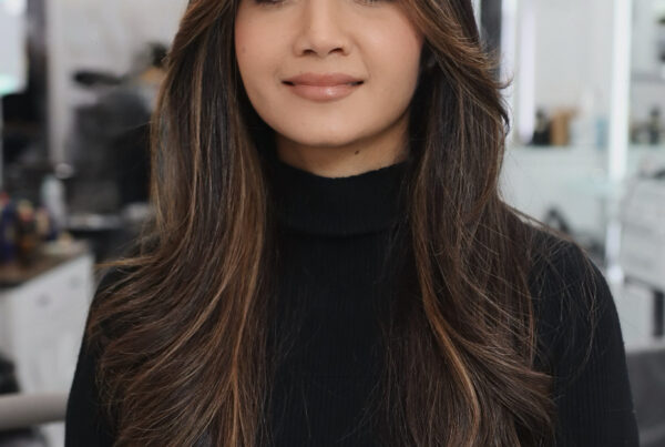 Smiling woman with long, layered brown hair and soft caramel highlights, wearing a black turtleneck inside Davida Salon in NYC.